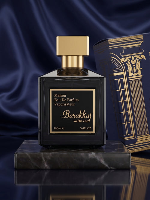 A dark glass perfume bottle with a gold cap and gold label reading 'Maison Eau De Parfum Barakkat satin oud 100ml' sits on a marble pedestal, with a matching ornate box beside it against a deep blue satin backdrop.


