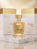 A perfume bottle labeled 'Barakkat gentle Gold' with a gold cap, displayed in front of two white and gold boxes on a marble surface against a warm beige background.

